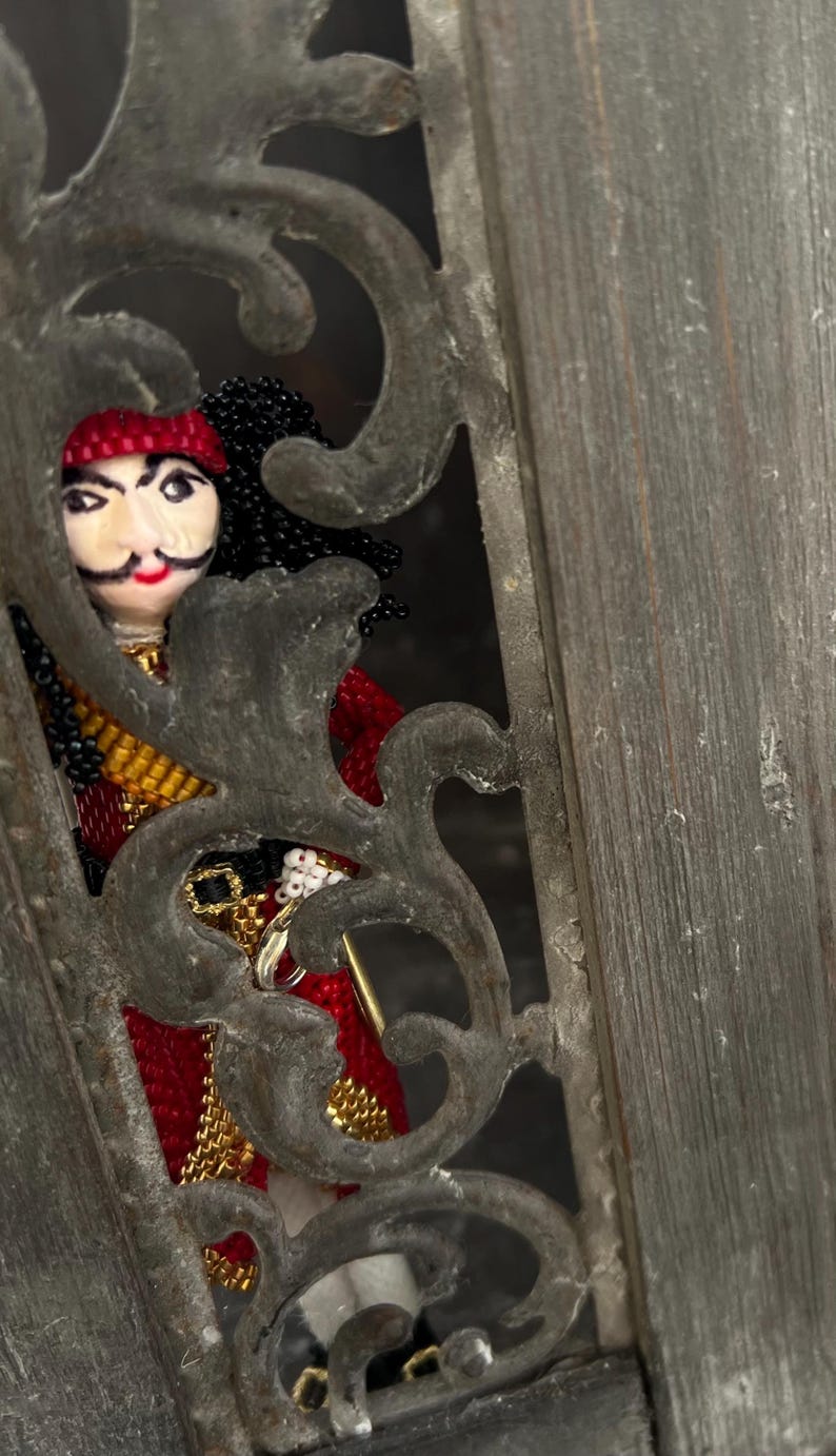 以下が含まれることがあります： 装飾的な金属製の枠から顔をのぞかせる、赤い帽子をかぶった黒髪のビーズ製の海賊人形。