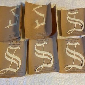 May include: Six individual cream-colored fabric letter S patches, each mounted on a brown paper backing. The paper features the printed text Reform Initials, Wash like Linen, Easy to Embroider, and instructions for creating monograms.