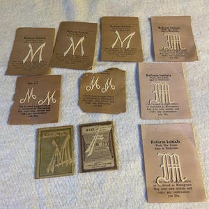 May include: A collection of nine vintage paper packets containing white embroidered initials. Each packet features the text Reform Initials, Wash like Linen, Easy to Embroider, and instructions for use. The letters M and F are visible in various fonts.
