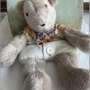 May include: A vintage-style teddy bear with a light brown plush body, button eyes, and a black stitched nose. The bear wears a cream-colored outfit with a button closure and a decorative collar with plaid accents. The bear is a classic toy.