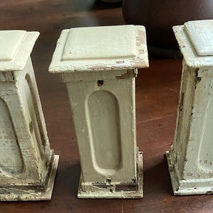 May include: Three antique, off-white wooden architectural elements with square tops and arched center panels. The paint is distressed, revealing the wood underneath. The pieces are arranged on a dark brown surface.