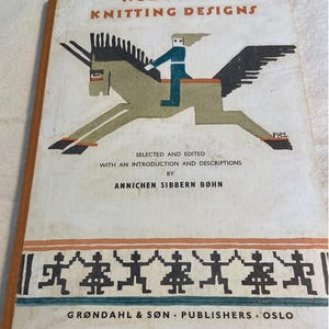 May include: A vintage book titled Norwegian Knitting Designs, selected and edited by Annichen Sibbern Bøhn. The cover features a stylized illustration of a figure riding a winged horse, with a border of dancing figures at the bottom, published by Grøndahl & Søn.