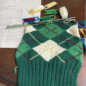 May include: A partially completed green, white, and yellow argyle knit project. The knitwear features a diamond pattern and is in progress with needles and yarn. A paper with instructions and yarn holders are also visible.