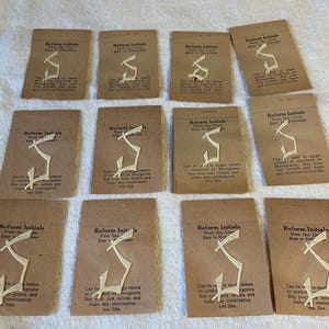 May include: Twelve individual tan paper packets labeled Reform Initials, each containing a white embroidered letter S. The text on each packet reads: Reform Initials, Wash like Linen, Easy to Embroider, Can be used in single letters or combined as Monograms.
