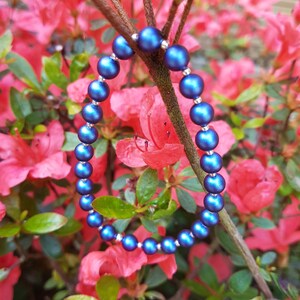 May include: A blue beaded bracelet with a metallic sheen, photographed against a backdrop of pink azalea flowers.