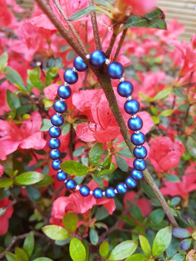 May include: A blue beaded bracelet with a metallic sheen, photographed against a backdrop of pink azalea flowers.