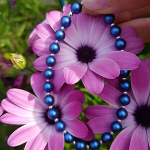May include: A blue pearl bracelet with a silver clasp, displayed against a backdrop of pink flowers.