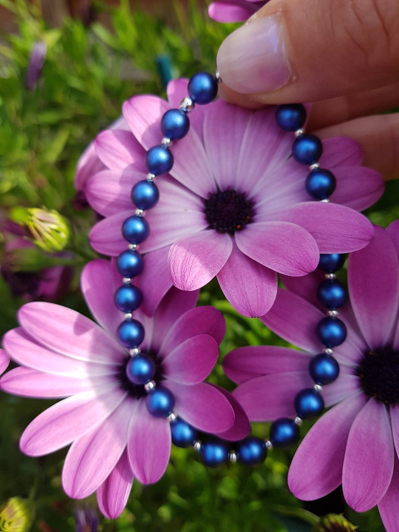 May include: A blue pearl bracelet with a silver clasp, displayed against a backdrop of pink flowers.