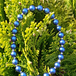 May include: A blue beaded bracelet with silver accents. The bracelet is made of round beads and is lying on a bed of green foliage.