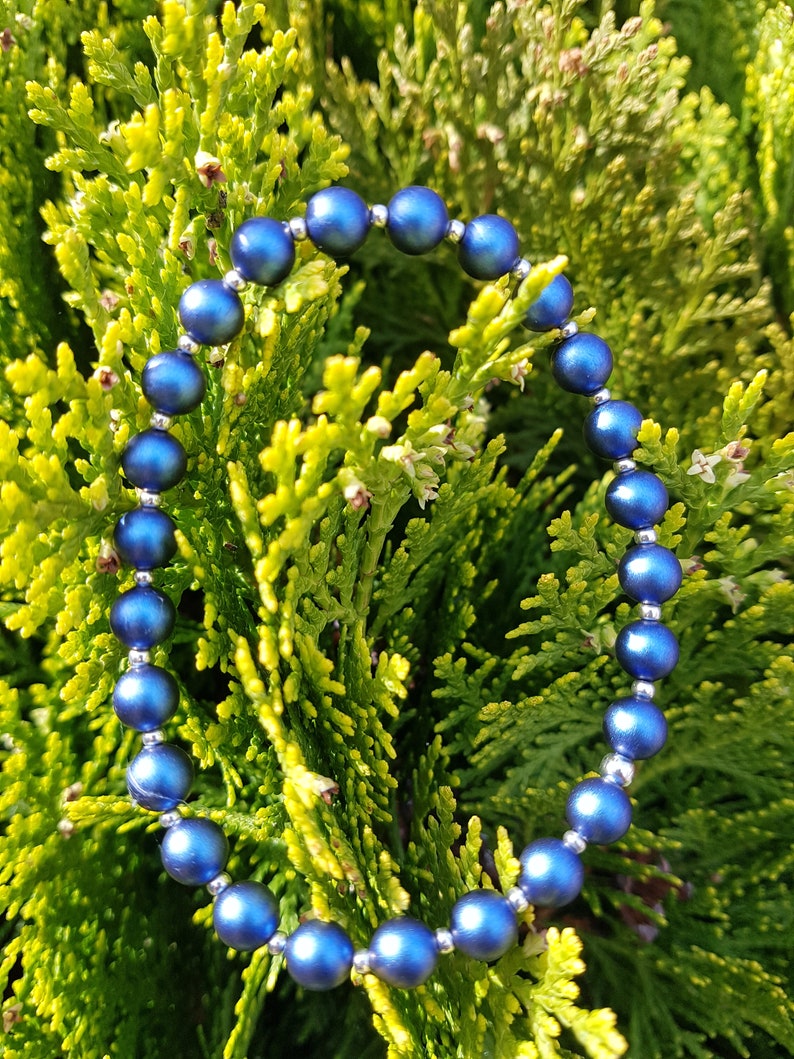 May include: A blue beaded bracelet with silver accents. The bracelet is made of round beads and is lying on a bed of green foliage.