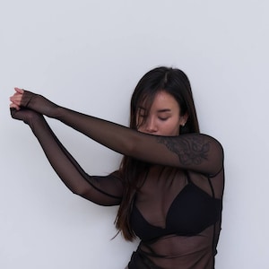 May include: A woman wearing a black sheer long-sleeved top over a black bra and brown leather trousers.