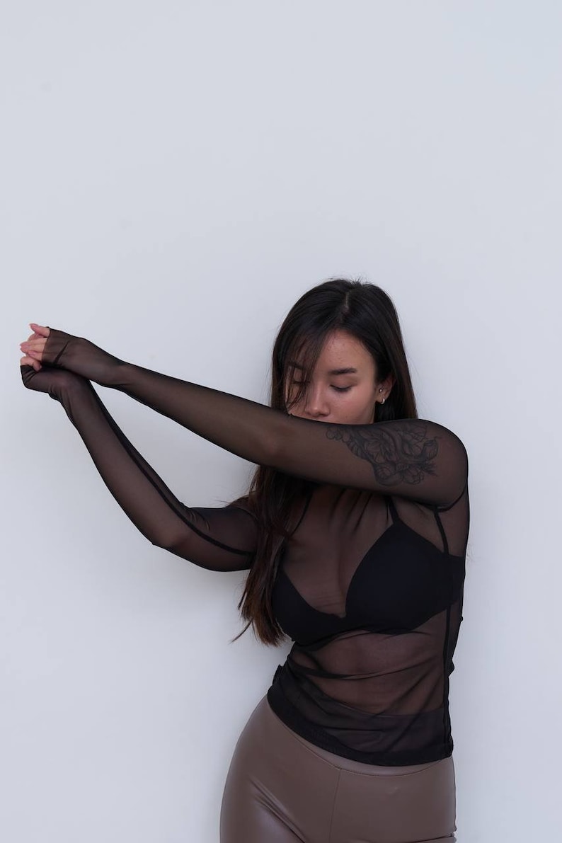 May include: A woman wearing a black sheer long-sleeved top over a black bra and brown leather trousers.