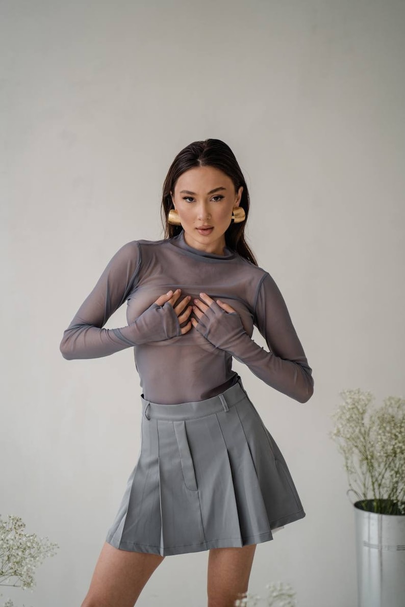 May include: A woman wearing a sheer grey long-sleeved top and a grey pleated skirt. She is wearing gold hoop earrings.