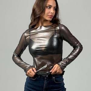 Pode incluir: Uma camisa de manga comprida prateada metálica com tecido transparente. A camisa é usada sobre um sutiã preto e combinada com jeans azul escuro.