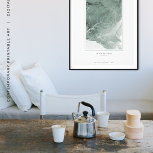 May include: A black framed print with a green abstract design and the text "ESTH&Eacute;TIQUE DE L'AVENTURE" and "LA VIE DE L'EAU". The print is hanging on a wall in a room with a wooden table, a teapot, and two cups.