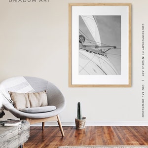 May include: A black and white photograph of a sailboat with the sail billowing in the wind. The photo is framed in a light wood frame and is hanging on a beige wall.