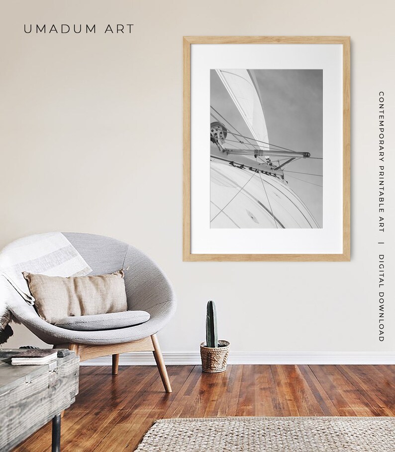 May include: A black and white photograph of a sailboat with the sail billowing in the wind. The photo is framed in a light wood frame and is hanging on a beige wall.