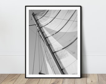 May include: Black and white photograph of a sailboat sail billowing in the wind. The sail is made of white fabric with black lines and is partially unfurled. The sail is attached to a mast and rigging.