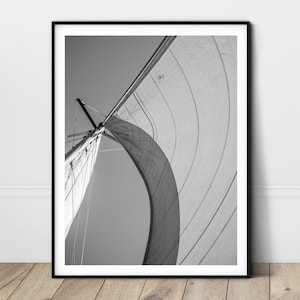 Op de afbeelding: Zwart-witfoto van een zeilbootzeil dat in de wind golft. Het zeil wordt van onderaf bekeken, met uitzicht op de mast en tuigage.