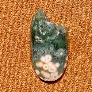 May include: A green and white ocean jasper cabochon with a unique pattern of white spots and swirls. The stone is oval shaped and has a smooth, polished surface.