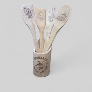 May include: A set of four wooden kitchen utensils with engraved designs, including a spoon, spatula, and fork, displayed in a cylindrical holder. The holder and utensils feature the Hogwarts School of Witchcraft and Wizardry logo and crests. The utensils are made of light-colored wood.