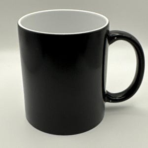 May include: A black ceramic mug with a white interior. The mug has a rounded handle and a cylindrical shape. The exterior is a solid, glossy black. The mug is designed for hot or cold beverages.