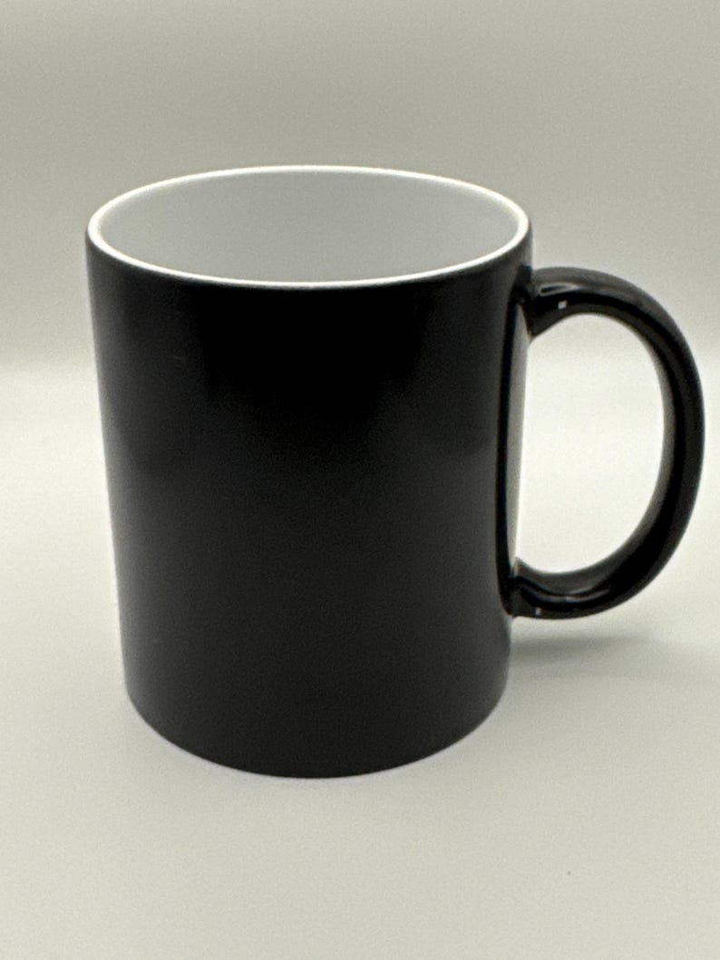 May include: A black ceramic mug with a white interior. The mug has a rounded handle and a cylindrical shape. The exterior is a solid, glossy black. The mug is designed for hot or cold beverages.