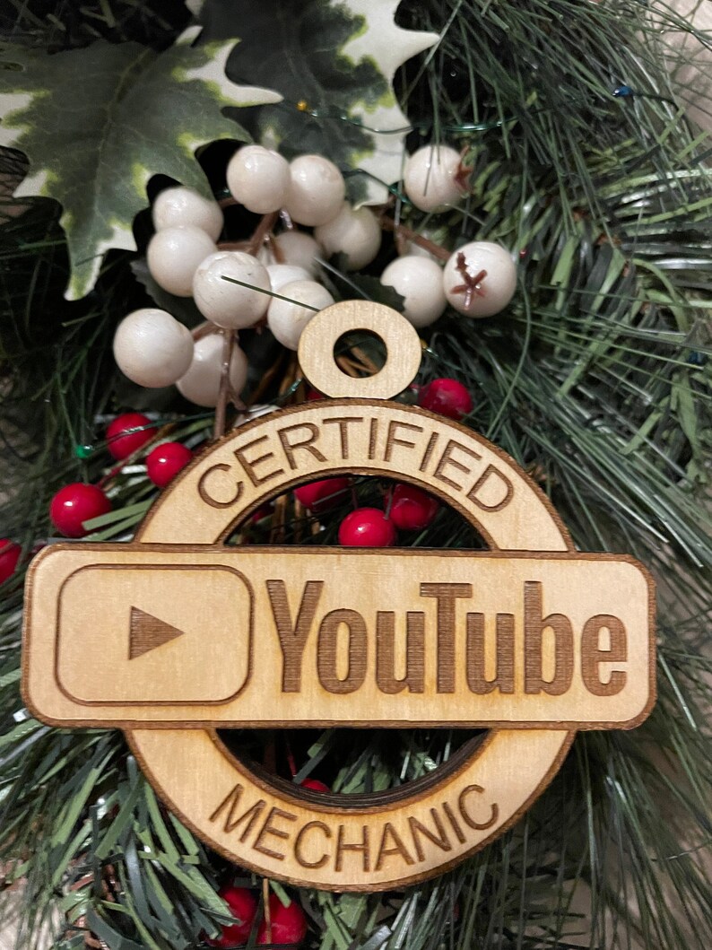 May include: A wooden Christmas ornament with the text "CERTIFIED YOUTUBE MECHANIC" and a YouTube play button icon. The ornament is surrounded by white and red berries.
