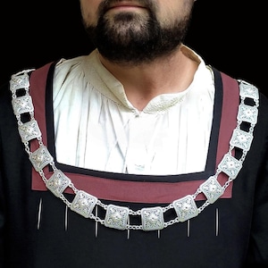 Può includere: Un uomo in un abito nero in stile Tudor, tra cui un cappello nero e una tunica nera con una sottoveste bianca. Una collana a catena d'argento con maglie quadrate e un colletto bordeaux accentuano il look. Lo sfondo è nero pieno.