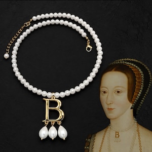 May include: A necklace featuring a strand of white spherical pearls with a gold-toned letter B pendant. Three teardrop-shaped white pearls hang from the bottom of the letter. The necklace includes a gold-toned metal chain extender and a lobster clasp.