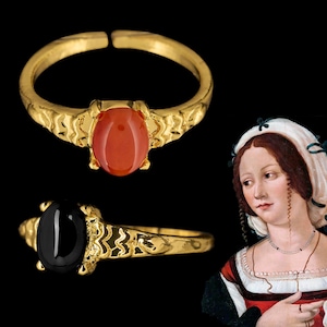 Peut inclure: Deux bagues dorées avec des anneaux texturés et des pierres cabochons ovales, l'une avec une pierre orange lisse et l'autre avec une pierre noire lisse, présentées à côté d'une peinture classique d'une personne portant une coiffe blanche.