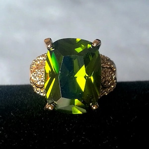 May include: A gold ring with a large, green, faceted gemstone. The ring has a decorative band with small, clear stones.