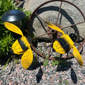 May include: Two metal bee garden ornaments featuring bright yellow bodies with black stripes and extended yellow wings. Each bee has a rounded black head and is mounted on a thin metal stake, positioned near a circular rusted metal wheel.