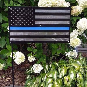 May include: A metal American flag with a blue stripe across the middle. The flag is mounted on a black metal pole and is displayed in a garden setting.