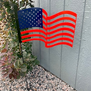 May include: A red, white, and blue metal American flag with a wavy design. The flag is attached to a black metal pole and is standing in a bed of small gray rocks.