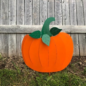 May include: A flat, orange metal pumpkin garden stake featuring a curved green stem and two green leaves. The pumpkin has vertical cutouts to define its shape and a thin metal spike at the bottom for insertion into the ground.