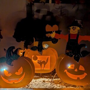 May include: Halloween-themed decorations featuring illuminated orange pumpkin cutouts with smiling faces and a scarecrow figure. The pumpkins have black leaf accents and a cat silhouette. The scene is lit with warm, inviting light.