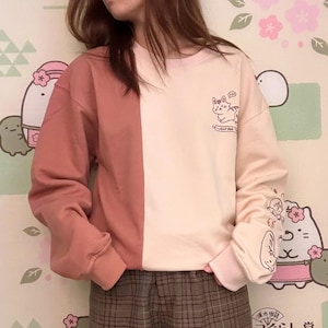 May include: A pink and peach colored sweatshirt with a graphic of a cat on the front. The sweatshirt has a split design with the left side being pink and the right side being peach. The cat graphic is wearing a pink headband and has a speech bubble that says "Meow". The sweatshirt has a white collar and cuffs. The sweatshirt is being worn by a person wearing brown plaid shorts.