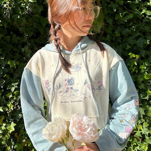 May include: A light blue and cream colored hoodie with a cartoon bear design. The hoodie has the text "Bearly Surviving" printed on the front. The model is holding two white peonies.