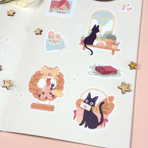 May include: A set of stickers featuring a black cat with a red bow, a house with a red roof, a window with a view of a cat sitting on a windowsill, a wreath with a cat and a girl, a cat carrying a letter in a basket, and a cat sitting on a windowsill. The stickers are on a white background with a pink border.