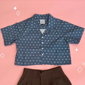 May include: A blue and white short-sleeved button-up shirt with a cat print. The shirt has a collar and a relaxed fit. The shirt is paired with a brown pair of pants.