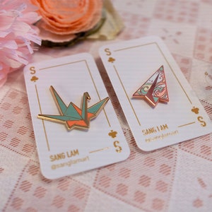 K&ouml;nnte beinhalten: Zwei Emaille-Pins auf wei&szlig;en Karten mit goldenen Akzenten. Die Pins sind beide Origami-Designs, eines ist ein Papierkranich in Gr&uuml;n- und Oranget&ouml;nen, das andere ist ein Papierflugzeug in Rosa- und Blaut&ouml;nen. Die Karten tragen den Text "Sang Lam" und "Sang I Am".