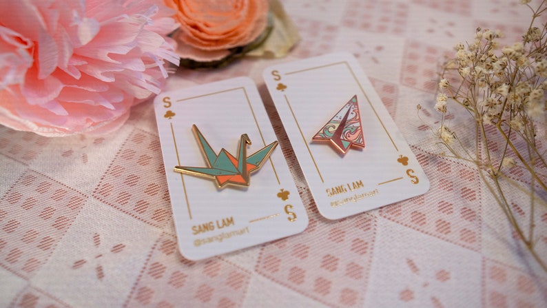 K&ouml;nnte beinhalten: Zwei Emaille-Pins auf wei&szlig;en Karten mit goldenen Akzenten. Die Pins sind beide Origami-Designs, eines ist ein Papierkranich in Gr&uuml;n- und Oranget&ouml;nen, das andere ist ein Papierflugzeug in Rosa- und Blaut&ouml;nen. Die Karten tragen den Text "Sang Lam" und "Sang I Am".