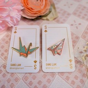 K&ouml;nnte beinhalten: Zwei Emaille-Pins mit Origami-Motiven. Der erste Pin ist ein gr&uuml;ner und orangefarbener Origami-Kranich. Der zweite Pin ist ein rosa und blaues Origami-Flugzeug. Beide Pins haben eine goldene Oberfl&auml;che und werden auf wei&szlig;en Karten mit dem Text "Sang Lam" und "@sanglamart" angezeigt.