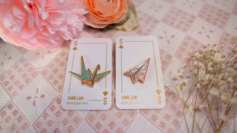 K&ouml;nnte beinhalten: Zwei Emaille-Pins mit Origami-Motiven. Der erste Pin ist ein gr&uuml;ner und orangefarbener Origami-Kranich. Der zweite Pin ist ein rosa und blaues Origami-Flugzeug. Beide Pins haben eine goldene Oberfl&auml;che und werden auf wei&szlig;en Karten mit dem Text "Sang Lam" und "@sanglamart" angezeigt.
