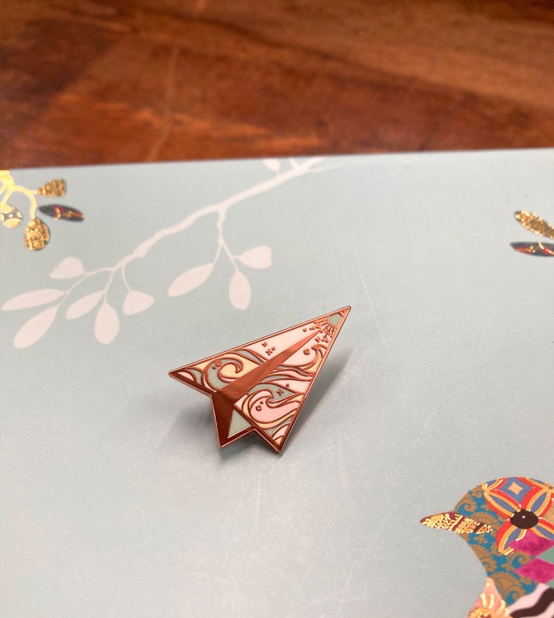 K&ouml;nnte beinhalten: Eine ros&eacute;goldene Papierflieger-Enamel-Anstecknadel mit wei&szlig;em Hintergrund und bunten Wirbeln. Die Anstecknadel befindet sich auf einem hellblauen Hintergrund mit einem wei&szlig;en Blumenmuster.