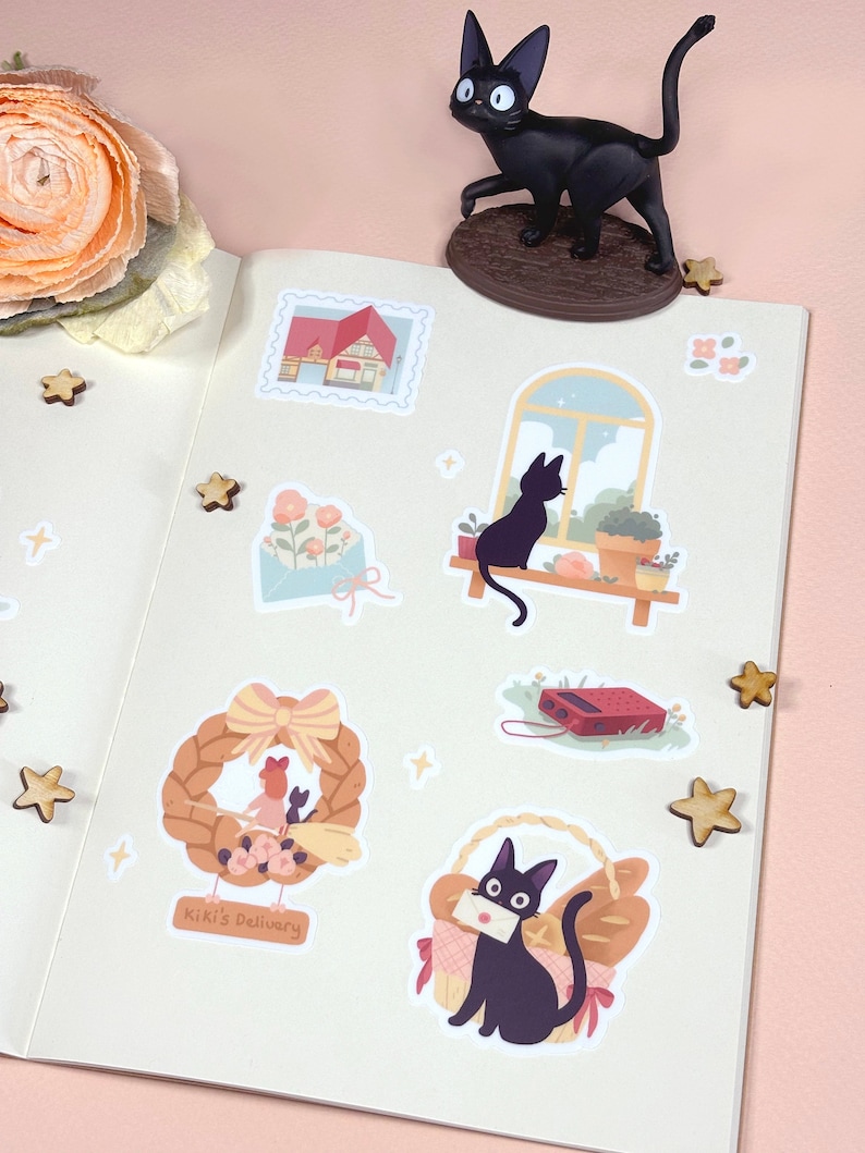 May include: A set of stickers featuring a black cat with a red bow, a house with a red roof, a window with a view of a cat sitting on a windowsill, a wreath with a cat and a girl, a cat carrying a letter in a basket, and a cat sitting on a windowsill. The stickers are on a white background with a pink border.