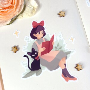May include: A sticker featuring a cartoon illustration of a girl with short black hair wearing a red bow and a pink dress, reading a book. A black cat with a red bow sits beside her. The sticker is surrounded by green leaves and white stars.