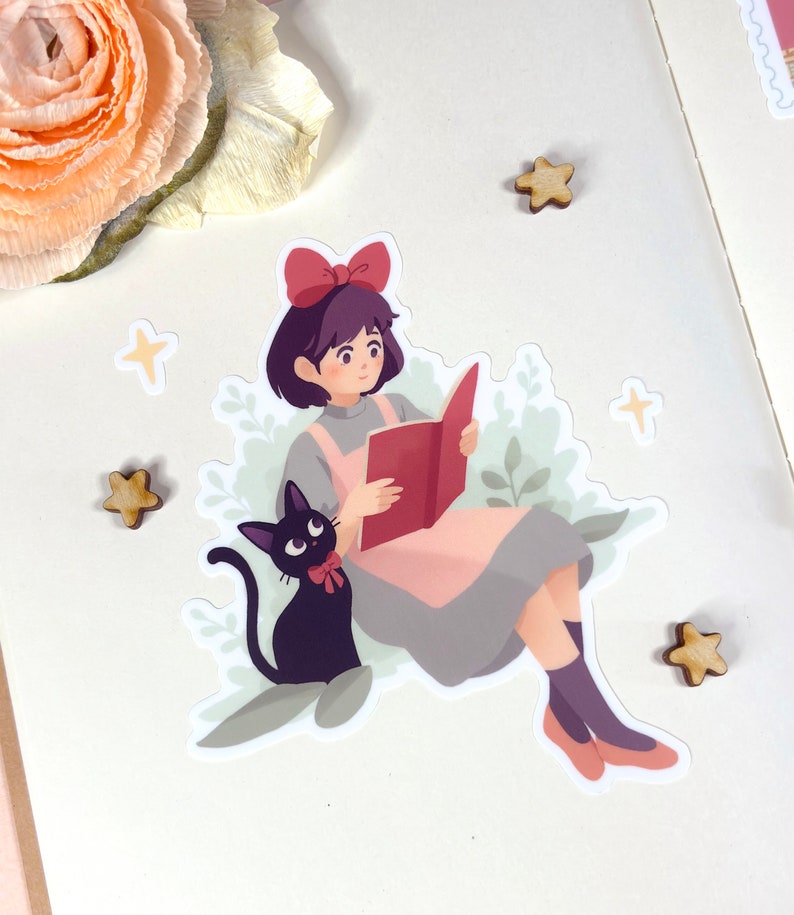 May include: A sticker featuring a cartoon illustration of a girl with short black hair wearing a red bow and a pink dress, reading a book. A black cat with a red bow sits beside her. The sticker is surrounded by green leaves and white stars.