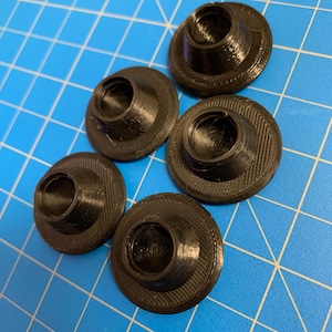 Op de afbeelding: Vijf zwarte ronde plastic doppen met een verhoogd midden. De doppen liggen op een blauwe achtergrond met een raster.